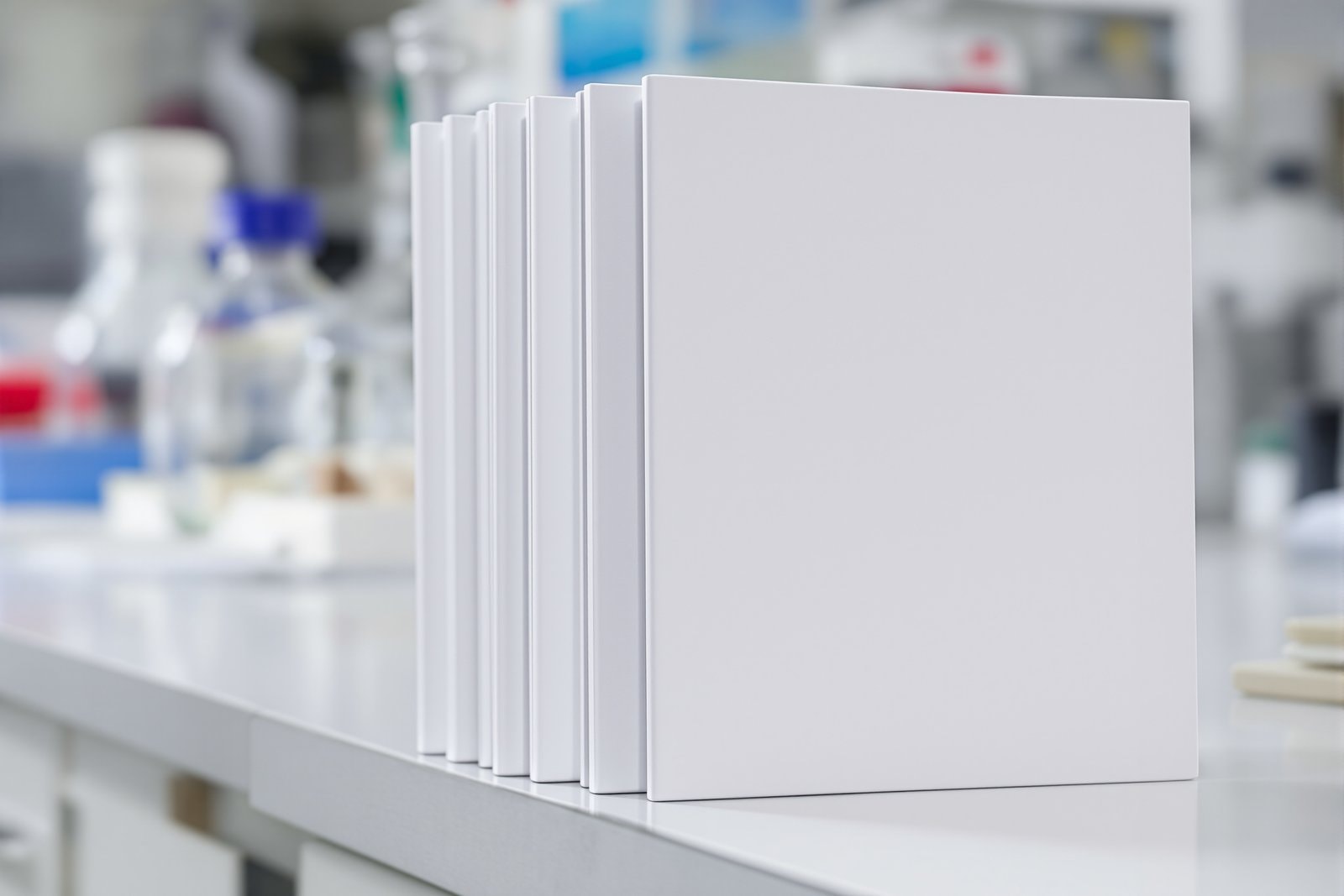 A stack of plain unlettered notebooks resting on the end of a laboratory bench with glassware blurred behind